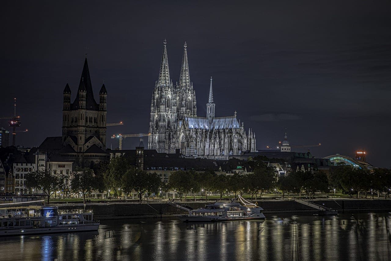 Gothic twin spires of Cologne Cathedral rising above the Rhine riverfront at dusk